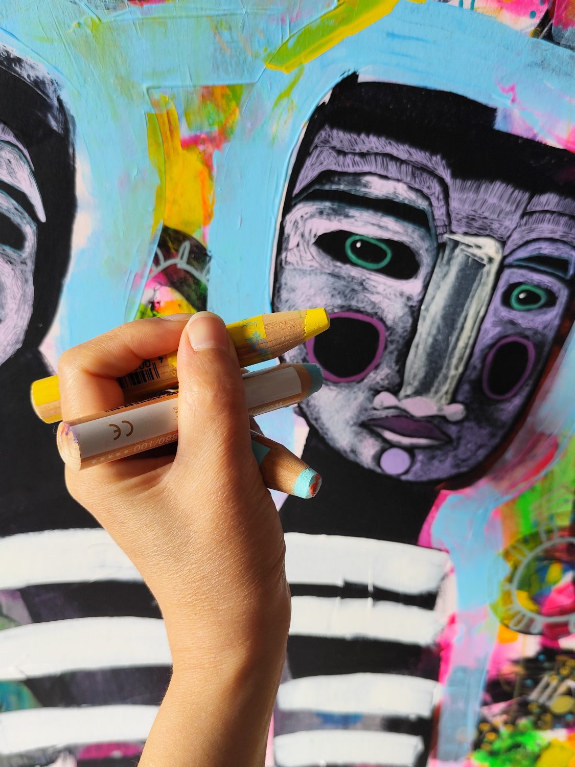 Eine Hand malt mit einem gelben Marker auf ein kunterbuntes Gesichtsgemälde.
A hand paints with markers a colorful portrait artwork